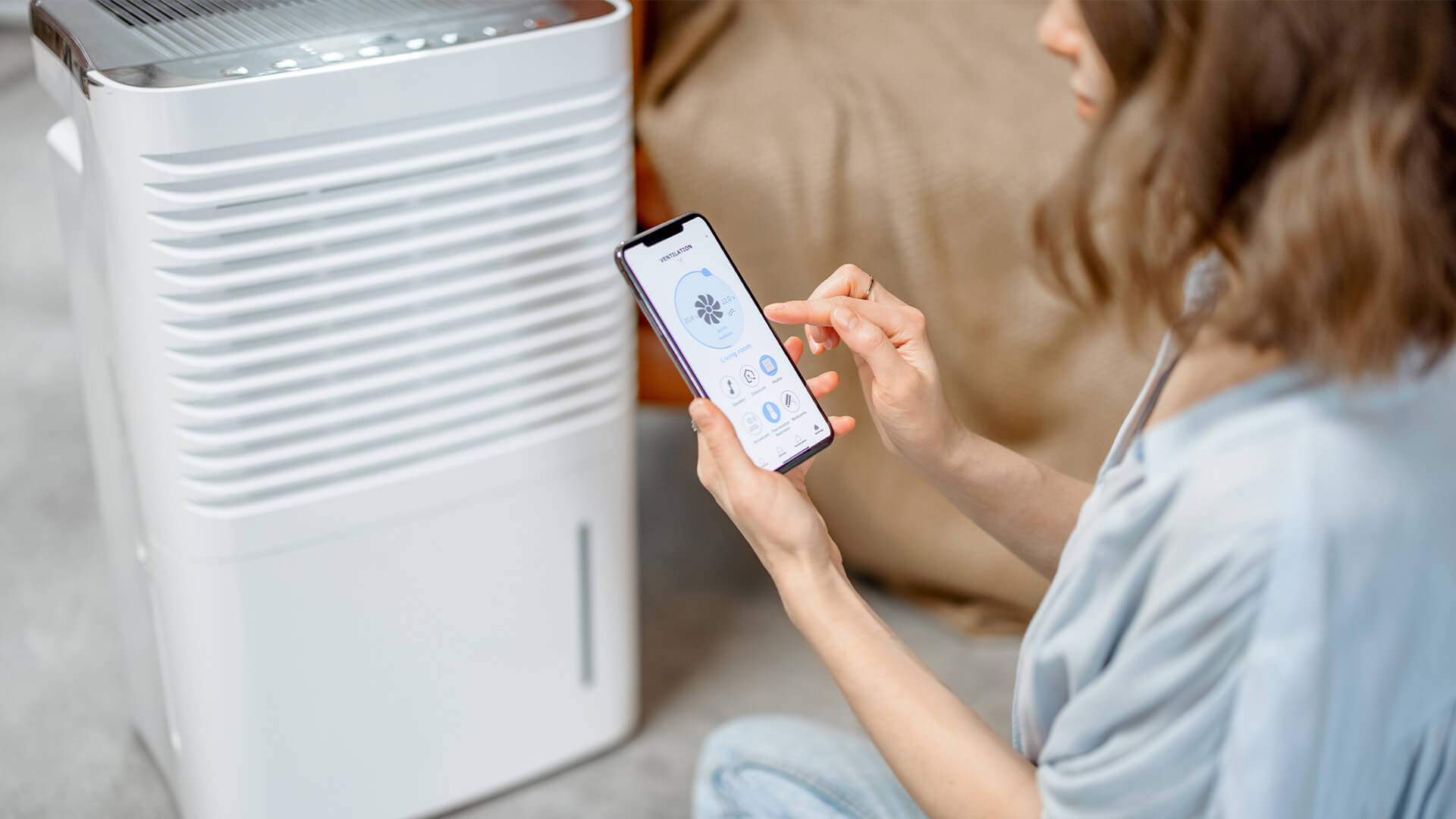 woman sitting near air purifier and moisturizer appliance near sofa monitoring air quality in phone.