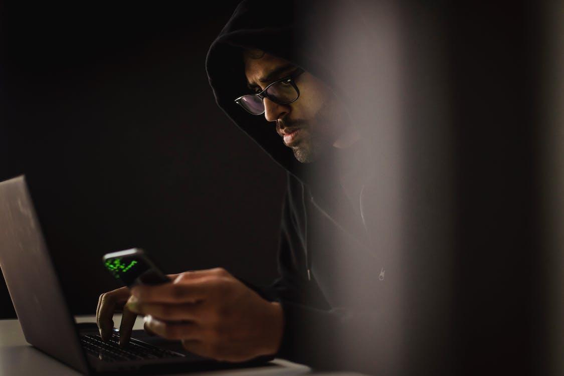Free Crop ethnic hacker with smartphone typing on laptop in darkness Stock Photo